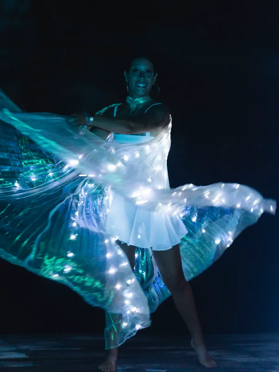 Bailarina girando con alas de luces LED azules y verdes sobre un escenario oscuro