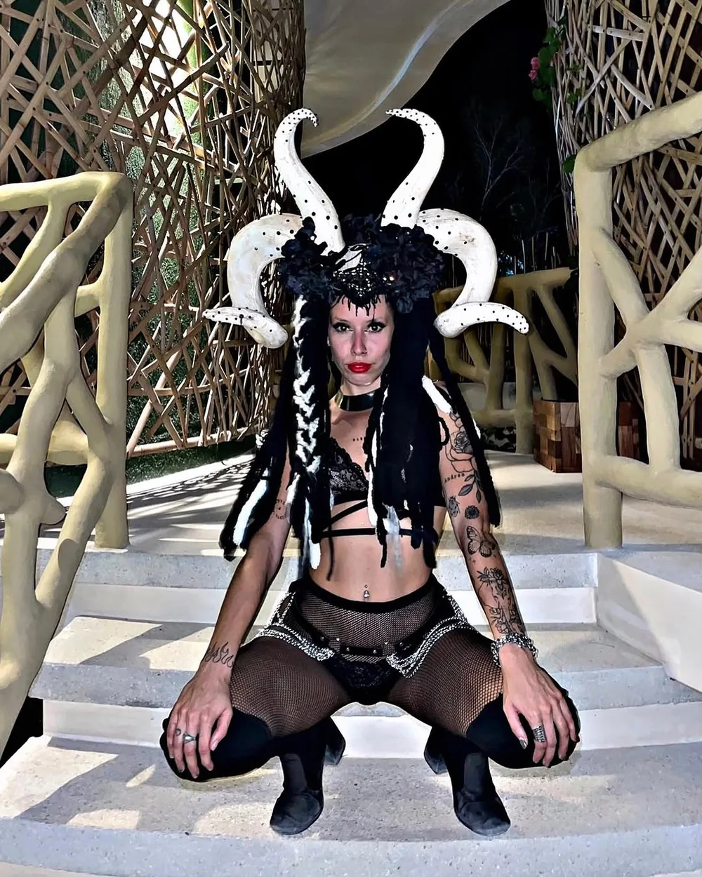 Performer con tocado de cuernos y vestuario negro, agachada en una escalera blanca